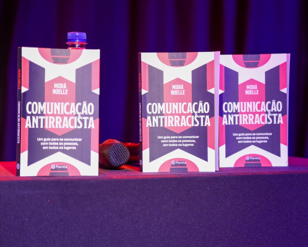 Três exemplares do livro Comunicação Antirracista, de Mídia Noelle, estão expostos em cima de uma mesa coberta por um tecido roxo escuro. As capas têm design gráfico nas cores vermelha, preta, branca e rosa, com o título em destaque. Entre os livros, há um microfone e uma garrafinha de água com tampa azul. Ao fundo, uma cortina roxa ilumina suavemente a cena.